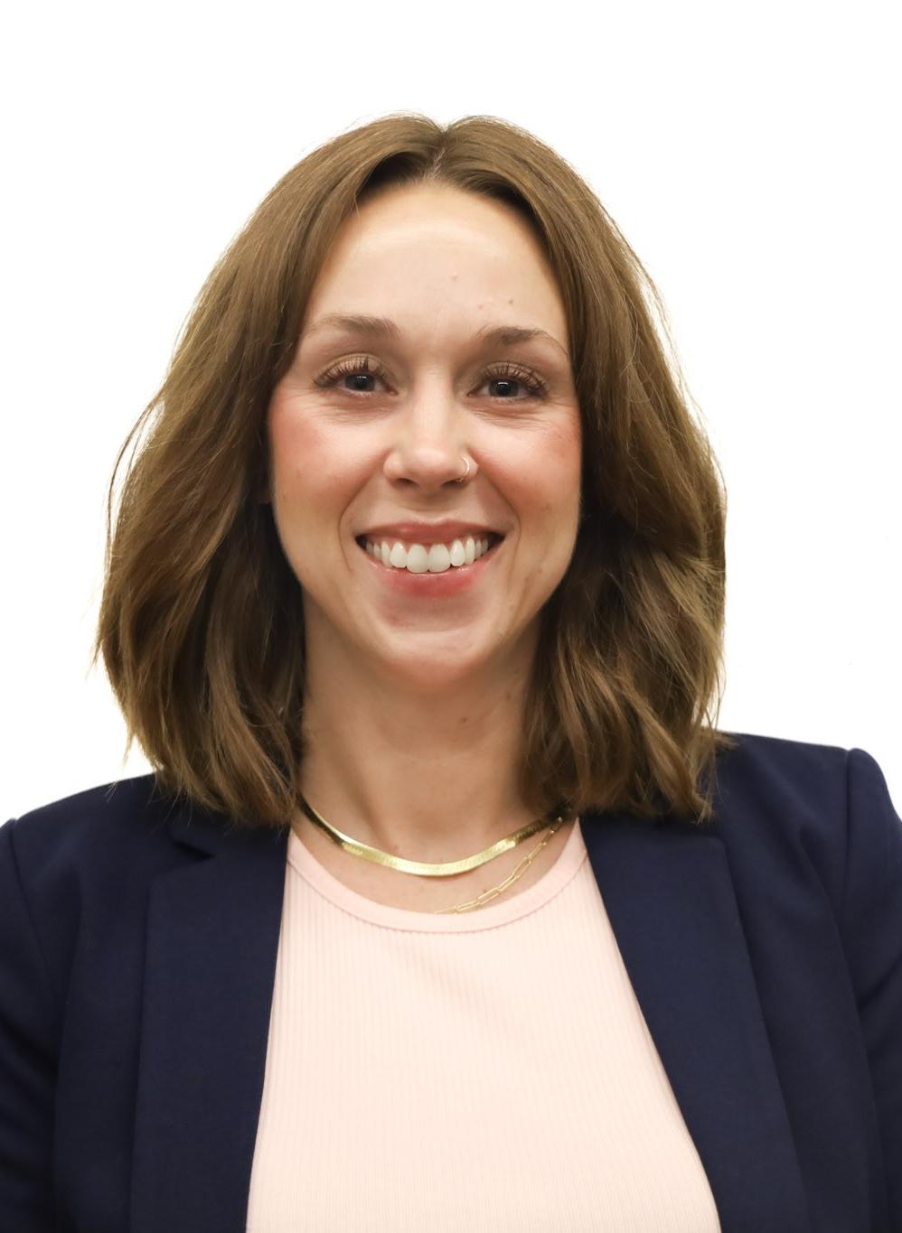 A white woman with shoulder length dark red hair wearing a blue jacket and pink shirt.